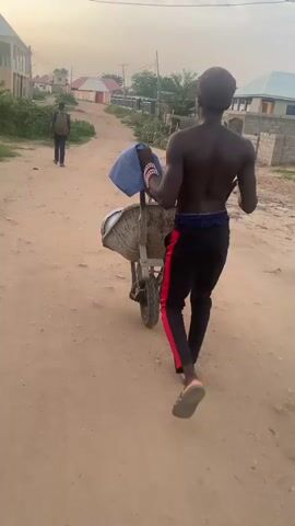 A young man with dark skin, wearing black pants with red stripes and sandals, pushes a wheelbarrow down a dirt road. He is shirtless and has a blue cloth and what appears to be beads in the wheelbarrow. He is walking away from the camera. In the distance, another person with a backpack is walking in the same direction. The environment is a rural or suburban area with buildings and vegetation lining the road. The sky is overcast with a warm hue, suggesting it might be dusk.