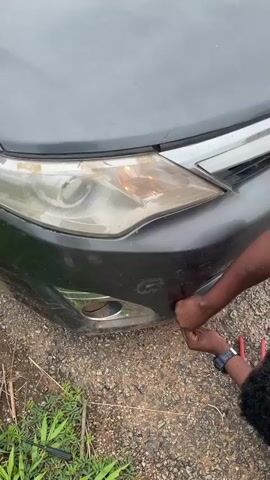 The video opens with a close-up of a dark-colored car's front bumper and headlight. A person's hands are visible, appearing to be working on the bumper. The scene then widens to show a man with dreadlocks wearing a patterned sweater and athletic pants standing next to the car, which is parked on the side of a road. He is looking at his phone. The setting is Oduape, Nigeria, with lush green vegetation on the side of the road and a wider landscape of trees and some distant buildings. A paved road stretches into the distance, with a few vehicles visible, including a truck and motorcycles. The sky is overcast with dramatic clouds, suggesting it might be late afternoon or a day with impending rain. The overall mood is somewhat somber or pensive, with the man engrossed in his phone and the car appearing to be either broken down or undergoing some minor repair. There is no visible text in the video.