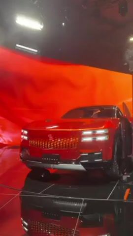 A red car on a black, reflective platform is displayed on a stage. The car has a flat, angular design, including rectangular headlights and a large, detailed grille. Behind the car is a large screen showing a swirling, abstract red pattern. The front passenger door opens up to the side. A bright light shines down from above.