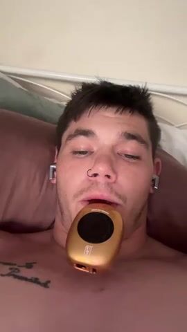 A person is lying in a bed, facing upward. They have short dark hair, a nose ring, and wear wireless earbuds. The person is shirtless, with a visible tattoo on their chest. They hold a small, gold-colored device in their mouth, which displays the words "Mexican Mango" and has a lighted circular interface. Vapor or smoke emerges from the device and the person's mouth. The bed has a white frame, brown pillow, and a green cushion. The background is a plain off-white wall.
