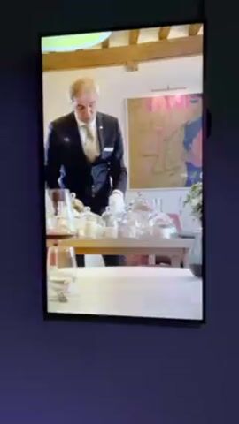 A screen shows a butler setting a table with desserts, each dessert under a small glass dome. The table is covered with a white tablecloth, and the man is wearing a suit and white gloves. The desserts include a dark liquid, a yellow cake, and thin biscuits.