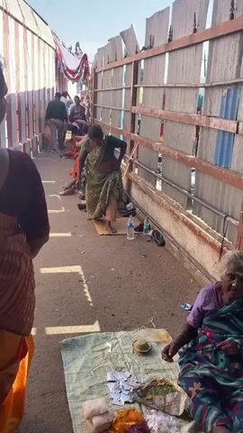 The video shows a walkway with a decorative canopy overhead. On the right side of the walkway is a corrugated metal fence. On the left side of the walkway are white and red striped pillars. Several people are seated or lying down along the right side of the walkway, next to the fence. A woman in a green sari is standing in the middle of the walkway, bending over. Several plastic water bottles and a pair of sandals are on the ground near her. In the foreground, a person with a purple and green shirt is sitting on a mat, with food and foil packets in front of them. Other people are walking away from the camera further up the walkway. The ground is paved and littered with small pieces of debris. The scene appears to be outdoors, in daylight, with a clear sky.