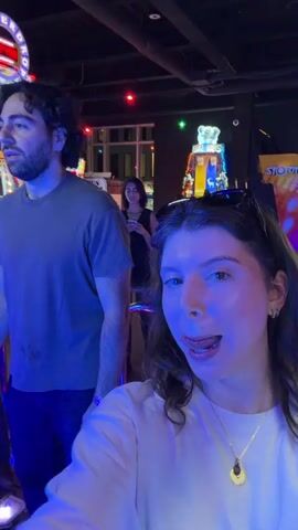 Two young men are playing a dance arcade game. They are both wearing casual clothing and are focused on the game's illuminated floor panels. The man in the foreground has dark, curly hair and is wearing a dark t-shirt. The man behind him has lighter brown hair and is wearing a white long-sleeved shirt. The game, labeled "MONSTERDINO," has a large screen displaying graphics and scores. The environment appears to be an arcade or entertainment venue, with other game machines and dim lighting, characterized by neon and LED lights. In the background, a woman in a black dress is looking at her phone. A woman's face is visible in the lower right corner, seemingly taking a selfie. The overall mood seems energetic and fun.