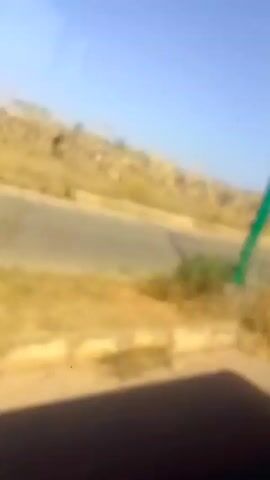 The video shows the view from a moving vehicle, looking out the side window. The scene is a rural or semi-rural landscape with a paved road running parallel to the vehicle.  The shoulder of the road is visible, with dry grass and some low-lying vegetation. On the other side of the road, there appears to be a field or open area with dry vegetation or crops. A few poles with green and white paint are visible along the road's edge. Further back, there are buildings, including a white building. The sky is clear and blue. A person walks on the shoulder of the road. The overall impression is of a drive through a sunny, somewhat arid area.