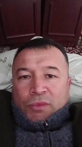 A man is speaking in the video, lying in bed. He is centered in the frame, wearing a zippered gray and blue turtleneck sweater under a dark jacket. His face is slightly puffy, and he is looking directly at the camera. Behind him is a pillow with faded lettering and a dark red wooden cabinet. The background is slightly out of focus. The man is touching his face near his ear at one point.