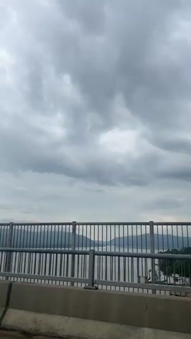 The video shows a view from a bridge looking out at a body of water with mountains in the distance. The sky is overcast with dramatic clouds. A metal railing runs along the edge of the bridge, and a concrete barrier is visible below it. The water appears calm with reflections of the sky and mountains. There are no people or vehicles visible in the shot.
