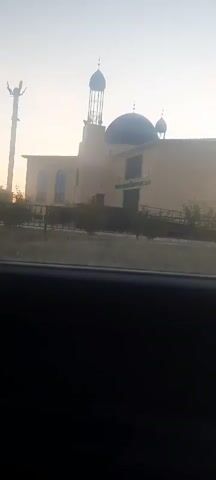 The video shows a mosque with a large blue dome and a tall minaret. The mosque is painted a pale yellow and has several windows. There is a fence in front of the mosque, and some trees and bushes are visible. The sky in the background is a pale orange, suggesting it is either sunrise or sunset. A utility pole with wires is visible to the left of the mosque. The video appears to have been filmed from inside a vehicle, as a dark interior of a car is visible in the foreground.