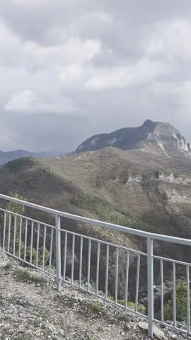 The video captures a panoramic view from a high vantage point, likely a mountain or hill, showcasing a vast natural landscape. The camera slowly pans from left to right, revealing rolling hills and distant mountains under a dynamic sky.

In the foreground, the ground is rocky and uneven, typical of a mountain peak or ridge. A light gray metal fence or railing runs along the edge of the viewpoint, serving as a safety barrier. The hillsides are covered with sparse vegetation, including some trees, suggesting a transitioning season or rugged terrain.

As the camera continues to pan, a Bulgarian flag mounted on a flagpole becomes visible. The flag, with its white, green, and red horizontal stripes, is gently waving in the wind, adding a touch of national identity to the scenic overlook. Beyond the immediate hills, a distant settlement or village can be discerned, nestled amidst the valleys.

The weather appears partly cloudy, with significant cloud cover but also patches of blue sky, indicating changing conditions. The presence of wind is evident from the waving flag. The scene conveys a sense of expansive natural beauty and tranquility from an elevated perspective. There are no people visible in the video, and no visible text is present within the frames. The location context indicates this is near Laki, Bulgaria, which aligns with the Bulgarian flag and mountainous terrain.
