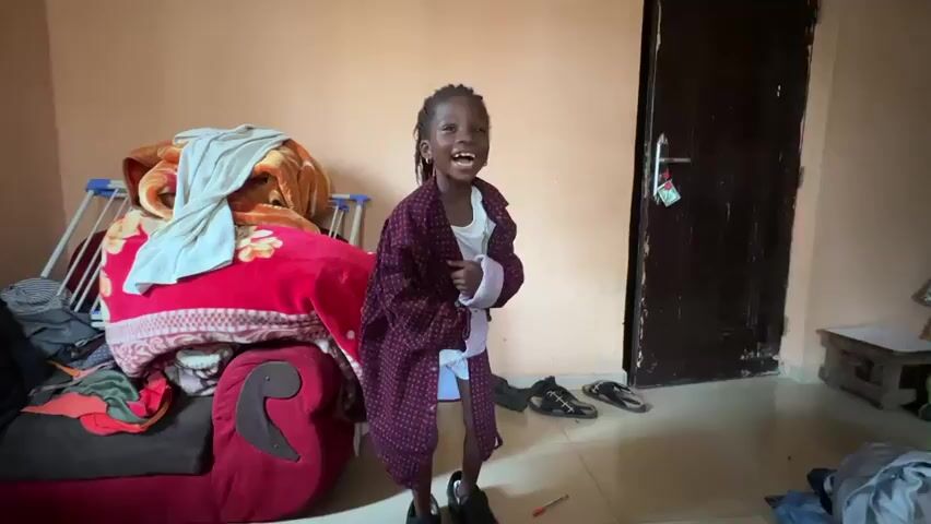 In a room with plain, light-colored walls, a young girl with dark skin and braided hair is the central subject. She is wearing an oversized maroon robe over a white garment and black shoes. Her expression is one of joy and excitement, with a wide smile and open mouth. She is standing in the middle of the room, turning her body as if dancing or posing.

To the left of the girl is a large, red sofa piled high with blankets and folded clothes. Behind the sofa, a blue laundry rack is visible. The floor is tiled, and several pairs of sandals and shoes are scattered around. A dark wooden door stands to the right of the girl, with a small piece of paper or tag hanging from its handle. In the background, there is a small wooden stool and some other items, including what appears to be a green container. The overall impression is that of a casual, domestic setting. The lighting suggests it is daytime, and there are no clear indicators of the weather. The atmosphere is lively and playful, primarily conveyed by the girl's exuberant demeanor. There is no visible text in the image.