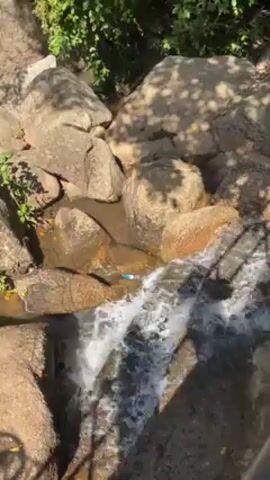 The video shows a small waterfall cascading over rocks. There are trees and shadows visible. The water appears clear and fresh. There is a blue plastic item stuck on a rock in the waterfall. A section of a metallic railing is visible at the bottom of the frame.