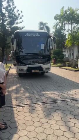 A bus with "PARIWISATA" and "AN TRANS" written on it is parked on a paved area. A person is standing on the left side of the frame, wearing a light-colored shirt and a dark skirt. The bus has a license plate that reads "B 7096 TZG". There are trees and buildings in the background. The lighting suggests it is daytime.