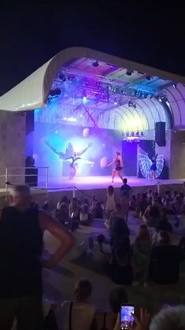 A nighttime outdoor performance is taking place on a stage under a curved white canopy. The stage is lit with vibrant purple and blue lights, and a large screen in the background displays a silhouette of a person with outstretched arms against a swirling blue and white pattern. Two aerial performers are suspended by red silks from above the stage; one is in the center, dressed in orange, and the other is on the right, wearing a butterfly-like costume. In front of the stage, a crowd of people, including many children, are seated on the ground, facing the performance. Some audience members are holding up their phones to record the event.