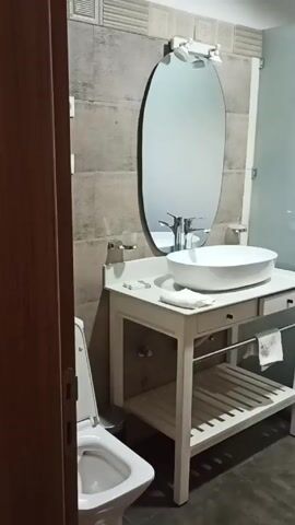 The video shows a bathroom. The toilet is on the left. There is an oval mirror above a sink on a white vanity. To the right of the sink is a frosted glass wall that separates the sink from the shower area. There are two small windows and a towel rack on the other side of the shower. The walls are tiled.