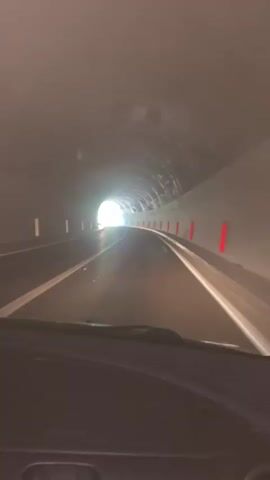 This video shows the perspective from inside a vehicle driving through a tunnel. The tunnel is illuminated by artificial lights and the bright light of the exit ahead. The road appears to be a two-lane highway with white and yellow lane markings. Red reflectors are visible on the tunnel walls at intervals. The tunnel walls themselves have a rough, possibly concrete texture. The view is from the driver's seat, showing the dashboard and windshield. The lighting suggests it could be daytime, as the exit is very bright, but the interior of the tunnel is somewhat dim. There is no visible text in the video. The scene conveys a sense of movement and progression towards the light at the end of the tunnel.