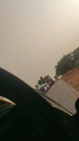 The video shows a car driving on a road with several trucks ahead. The sky is overcast. The road is narrow, bordered by trees and a reddish-brown shoulder. The view is partially obstructed by the car's frame. In the driver's side mirror, a hand with a bracelet can be seen.