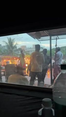 The video, filmed from inside looking out through a window, shows a group of people gathered on an outdoor patio or terrace during the early evening or dusk. There are approximately six to seven individuals visible. In the center, a woman wearing a sari appears to be leading an activity, possibly a religious ritual or ceremony, as she faces a collection of lit candles or small lamps arranged on the patio floor. Other individuals, mostly men in casual attire, stand around her, observing or participating. One person is seated in the foreground, viewed from behind, also looking towards the activity.

The patio surface appears wet, suggesting recent rain, and there are water droplets on the windowpane, blurring the view slightly. String lights are strung across the patio, adding to the evening ambiance, and potted plants are visible. In the background, typical urban buildings and palm trees can be seen under a soft, cloudy sky. Bottles, possibly water or soda, are partially visible in the immediate foreground, inside the room from where the video is shot. The overall scene suggests a calm, communal gathering, likely a special event or prayer, in an urban setting like Chennai, India. No discernible text is visible.