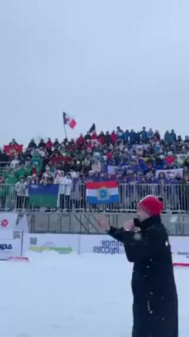The video shows a person, presumably the emcee, throwing a snowball at a crowd in a snowy stadium.  The person is wearing a black coat with "УДМУРТИЯ" printed on the back. They have their back to the camera and are facing the crowd, who are sitting in bleachers and holding various flags. Snow is falling during the event.  There are advertising banners around the field.