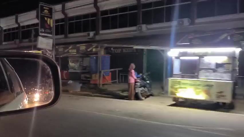 The video shows a street scene at night, observed from inside a car looking through the side mirror. The street is lined with buildings, most of which have closed storefronts. Several people are seen moving about, some on motorcycles, and others standing in front of the shops. There are several street vendors as well. The general atmosphere is calm and quiet, with low-level illumination from the streetlights and storefronts. The footage appears to be raw and unedited.