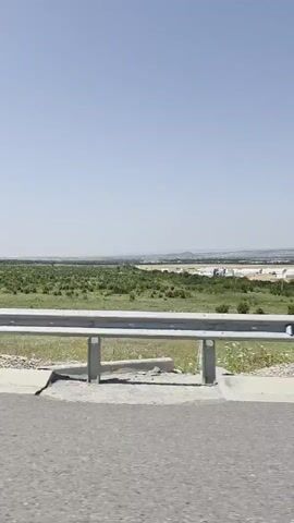 A video captures a view from inside a moving vehicle. The camera is positioned to show the road ahead and the surrounding landscape. A guardrail runs alongside the road, separating it from a field of green vegetation. In the distance, a hazy horizon suggests more land or possibly a city. The sky is clear and blue, indicating a sunny day. A side mirror reflects a person holding a phone, presumably the person filming. The vehicle appears to be on a highway or a similar paved road. The footage is characterized by the motion blur typical of a moving vehicle, with the landscape shifting and changing as the car progresses.