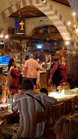 In a dimly lit restaurant with stone arched ceilings, patrons are seated at wooden tables. In the background, a performance is taking place. Two women in traditional red and black flamenco dresses with red flowers in their hair are clapping and dancing. A man in a red shirt, black vest, and red beret is also dancing and gesturing with his hands, interacting with the dancers and the audience. Another man in a pink shirt is standing near a bar area, possibly part of the performance. Several other people are observing the performance. The restaurant appears to be decorated with rustic, traditional elements, including pots, pans, and shelving filled with various items.