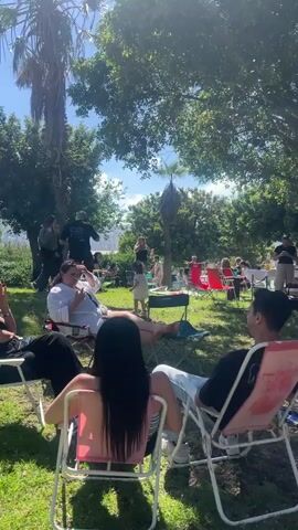 The video shows a daytime outdoor scene with a group of people gathered in a park or picnic area. 

Initially, the camera focuses on two individuals sitting in lawn chairs, then pans across the area to reveal a larger group, some sitting and some standing, engaged in various activities. There are tables set up with food and drinks, suggesting a picnic or barbecue.

The camera moves past tables, trees, and people, eventually showing individuals in orange vests standing near a grill. The scene pans further, revealing a parking lot filled with sailboats. Finally, a woman with pigtails walks towards the sailboats. The sky is clear and blue.