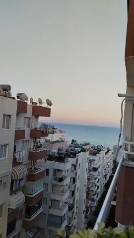 This video shows a view from a balcony overlooking a coastal city, likely Mezitli, Turkey. The scene is dominated by a row of multi-story apartment buildings with many balconies, some adorned with satellite dishes and solar water heaters. Laundry hangs from some of the balconies, suggesting daily life is in progress. Beyond the buildings, the vast expanse of the sea stretches to the horizon. The sky is a clear, pale blue, transitioning to soft hues of pink and orange near the horizon, indicating either sunrise or sunset. A small, pale moon is visible in the sky. The overall mood is peaceful and serene, capturing a tranquil moment in a densely populated urban environment by the sea. There is no visible text in the video.