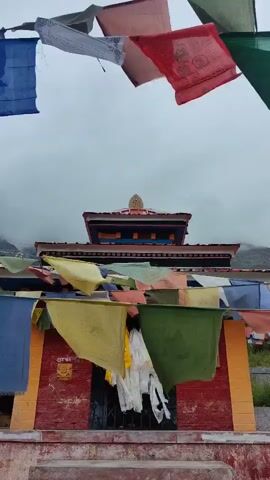 The video shows prayer flags in front of a temple. The flags are colorful and have different patterns on them. They are strung across the front of the temple, fluttering in the wind. The temple itself is made of red brick and has a traditional architectural style. The sky is overcast and the overall mood is peaceful.