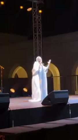 A woman wearing a white hijab and a long flowing white dress is singing on a stage. She is holding a microphone in her right hand and gesturing with her left hand. The stage has two speakers on either side of her. Behind the stage is a dark wall with arched openings lit in yellow. There is also a metal lighting rig behind her. The overall lighting is bright.