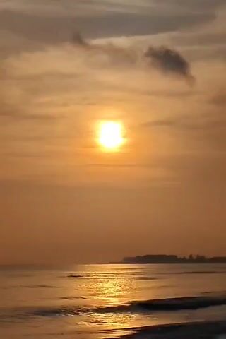 The video shows a scenic view of the ocean at sunset. The sun, a bright disc in the sky, casts a golden glow over the water and sand. Several clouds of varying shapes and sizes are scattered across the sky, some darker than others. The waves gently roll onto the beach, reflecting the sunlight. A strip of land with trees is visible in the distance. The camera appears to be stationary, capturing the serene and tranquil scene.