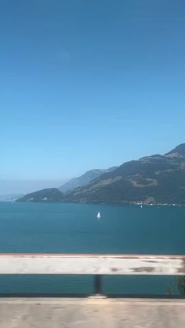 This is a side-view, somewhat blurry video of a body of water, likely a lake, under a clear blue sky. Mountains covered in green vegetation rise from the far shore. A white sailboat is visible on the water. In the foreground, there is a blurry railing, possibly from a bridge or walkway. The camera appears to be moving.