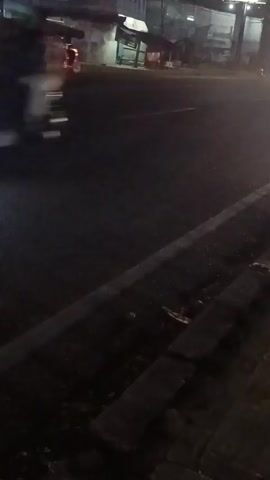 The video shows a nighttime view of a street. In the distance, a red vehicle with flashing lights is visible. A person on a scooter with blue lights passes by in the foreground, followed by another motorcycle with red taillights. A white truck is also seen driving on the street. The environment appears to be urban with buildings in the background. The ground is wet, suggesting it might have rained.