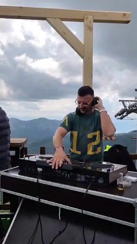 A man wearing a green and yellow t-shirt and headphones is DJing at an outdoor event. He is standing behind a DJ setup, adjusting knobs and sliders. Another man in a black vest and baseball cap is standing to the left, looking down at his phone. In the background, there are mountains and a cloudy sky. A wooden sign hangs above them that reads "2035 ABOVE THE CLOUDS".