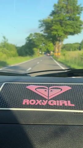 A first-person perspective video shows a car interior, focusing on a RoxyGirl branded mat on the dashboard. Through the windshield, a road is visible, bordered by green trees and fields under a clear blue sky. Another car is in the distance on the road. The camera is stationary.