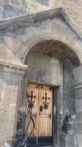 A vertical video shows a stone building, possibly a church, with a large arched entrance and wooden doors. The stone walls are weathered and feature decorative carvings, including crosses and intricate patterns. The building appears to be ancient. The camera pans up the facade, revealing arched windows and a dome structure at the top. The sky is blue with some white clouds.