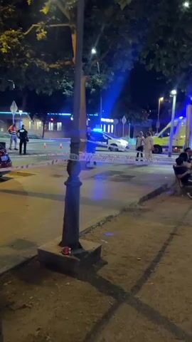 The video shows a scene at night that appears to be the site of an emergency or police incident. The area is illuminated by streetlights and the flashing blue lights of emergency vehicles, including an ambulance and police cars. People are present, some appearing to be bystanders or onlookers, while others are emergency personnel. There is caution tape cordoning off a section of the street, and several people are gathered near the tape, including individuals in what appear to be law enforcement or emergency services uniforms. In the foreground, a group of people, including children, are sitting on a bench. The environment includes trees, sidewalks, and a lamppost.