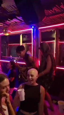 The video captures a lively scene inside a nightclub filled with people dancing and enjoying the music. The club is decorated with neon lights, a disco ball, and what appears to be a thatched roof or bamboo-style design feature. The color scheme is predominantly dark, with vibrant reds, blues, and purples from the lighting creating a dynamic ambiance.

A saxophone player is performing in front of a mirror or window framed with bright red neon lights. People are standing around him, some are dancing, and others are holding drinks. Several people are visible up-close, including a woman with a shaved head holding a white cup, a person in sunglasses wearing a hooded vest, and a couple of girls with their friends who are happily dancing.

The camera moves, showing more of the crowd and a neon sign that reads "Tequila Sundays". The overall atmosphere appears festive and energetic, with people enjoying the nightlife and music.