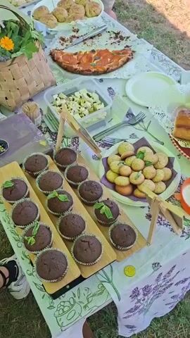 This is a close-up, top-down, slightly tilted shot of a table laden with various food items, presumably set up outdoors. In the foreground, on the left, are rows of chocolate cupcakes topped with green leaves. To the right of the cupcakes is a white plate filled with golden-brown cookies, also garnished with green leaves. Further back on the table, a pizza with visible toppings and a rectangular container of cubed white cheese are visible. To the left of the pizza, a woven basket overflowing with flowers, including orange and yellow blooms, is present. The table is covered with a tablecloth featuring a lavender print. Two people are partially visible in the frame; one on the left has their legs and feet, wearing sneakers, in the lower left corner, and another on the right with their lower torso and legs, wearing shorts, at the top right. The overall impression is of an outdoor picnic or market stall display.