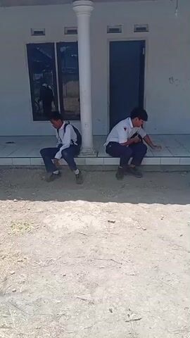 Two young males wearing school uniforms are outside a building. One male is sitting on a raised platform, leaning against a pillar, facing left. The second male is crouching on the platform, facing right. A motorcycle is parked in front of the building. The second male stands up, grabs a bag, and walks toward the motorcycle. The first male remains seated. The second male gets on the motorcycle and drives away, turning left. The first male remains seated. The video ends.