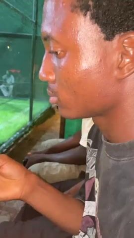 A medium shot of two young men sitting next to each other. The man on the left is wearing a white t-shirt and beige shorts, and he is holding a black controller. The man on the right is wearing a black t-shirt and is looking to his left. In the background, there is a green net, a grassy area, and several other people. The scene appears to be outdoors at night.