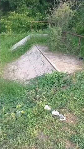 The video shows a concrete ramp structure overgrown with grass and vegetation. In the background, there are trees and a blue sky with some clouds. There is also some trash visible in the foreground. The camera is moving and appears to be handheld, with occasional shaky movements. The audio consists of indistinct speech, possibly a conversation.
