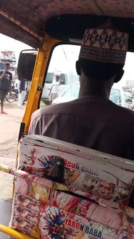 The video shows a ride in a yellow motorized tricycle, commonly known as a keke or rickshaw, through what appears to be a crowded market area in Nigeria. The view is from inside the vehicle, behind the driver. The driver is wearing a brown shirt and a patterned kufi cap. The back of the driver's seat is decorated with images and the words "Yaron Baba." Outside, people are walking and motorcycles are moving in the street. Some individuals are carrying items on their heads. There is a white van with a cracked windshield visible in the background. The road is unpaved and dusty. The keke moves slowly through the congestion. The sound of the keke's horn is heard frequently.