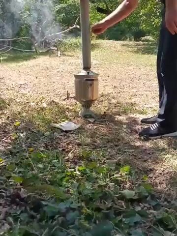 A person in black pants and a dark shirt is standing outdoors, near a smoking apparatus that appears to be a samovar. The person is holding a white object, possibly a cigarette or a piece of kindling, and reaching towards the top of the apparatus, where smoke is emanating. The ground is covered with dry leaves and some green vegetation. In the background, there are trees and branches, suggesting a wooded or natural area. The lighting indicates it is daytime.