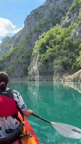A person is kayaking on a serene, turquoise lake, paddling through a narrow canyon with towering, lush green mountains on either side. The sun is shining brightly, indicating it's likely midday, and the sky is a clear blue with a few fluffy white clouds. The water is calm and reflects the steep, rocky cliffs and the vegetation clinging to them. The person is wearing a red life vest over a white shirt with a blue and black pattern. They are holding a white kayak paddle with a black shaft. The kayak itself is orange and red. The overall mood is peaceful and adventurous, showcasing the beauty of nature in this stunning canyon. No visible text is present on the objects or scene.