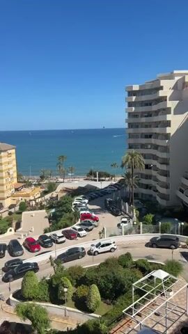 The video presents a view from a high vantage point overlooking a coastal scene. Below, there is a parking lot filled with cars, a road leading to the beach, and a strip of sandy beach. Palm trees dot the landscape. The ocean is a vibrant blue. In the distance, a parasail with two visible occupants is suspended above the water. A boat is also seen pulling the parasail. The sky is clear and blue.  A tall white building is partially visible on the right side of the frame.