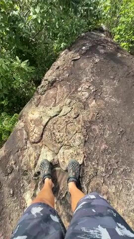 The video begins with a low-angle shot of a person's feet, clad in black sandals, standing on a large, rough, grey rock. The rock stretches out ahead, covered in textured patterns and cracks. Lush green foliage borders the left and right sides of the rock. The camera then pans up and to the right, revealing a breathtaking panoramic view. The perspective shifts to a higher vantage point, looking out over a vast, verdant landscape. Rolling hills covered in dense green trees stretch as far as the eye can see. In the distance, several large, distinct rock formations rise dramatically from the landscape, some with their peaks shrouded in wispy white clouds. The sky above is a bright blue, dotted with scattered white clouds, and the sun is visible, casting a bright light. The camera continues to pan, showing more of the stunning natural scenery and the interplay of light and shadow on the mountains and valleys below.