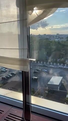 The video shows a view from a window overlooking a city during what appears to be late afternoon or early evening. The sun is shining brightly, casting a glare on the window and creating lens flare effects. In the foreground, a white Roman shade is visible, pulled halfway down, with a bead chain hanging to the left. Through the window, a parking lot filled with cars is seen, along with some buildings and trees. The cityscape extends to the horizon, with more buildings and greenery visible. The sky is blue with scattered clouds. The camera pans slightly to the right, revealing more of the city and its architecture, including some older, ornate buildings with light pink facades and red roofs. The reflection of a person taking the video is visible on the window in the middle section of the frame.