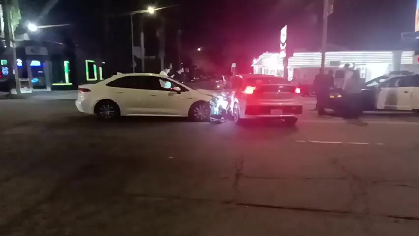 At night, two white cars, one appearing to be a Toyota Corolla and the other possibly a Hyundai Ioniq, are situated with their front ends smashed together, indicating a collision. Debris lit by headlights is visible on the road. Several people are standing nearby, and a police car is present. A police officer is standing near the police vehicle. Buildings with lighted signs are visible in the background, and a billboard displays a burger advertisement.