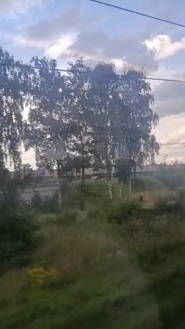 The video shows a view from a moving vehicle, likely a train, looking out at a landscape. The time stamp indicates the video is from a few seconds duration. The scene features a bright sky with scattered clouds, trees, and vegetation. There appears to be a railway line with graffiti on a barrier or wall beside it. Power lines are visible in the foreground and across the sky. The overall impression is a suburban or rural environment with signs of human activity. There are no people or animals visible in the footage.