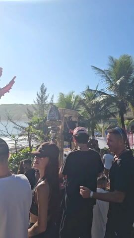 The video shows a beach party with a DJ performing on stage, surrounded by a crowd of people. The stage is decorated with a large red bird sculpture and green foliage. The sea is visible in the background, with waves breaking on the shore. The sky is blue with scattered clouds. The sun is shining brightly, creating a glare in some shots. People in the crowd are wearing casual clothes and hats. Some are dancing and talking to each other. The camera moves around to show different angles of the scene.