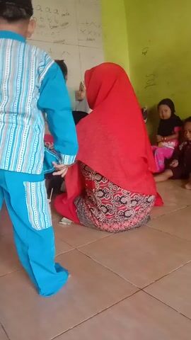 A teacher wearing a red hijab and a patterned dress kneels on the floor, interacting with several young children who are also on the floor. A whiteboard is visible on the left, with some drawings and what appear to be letters and numbers. The children are arranged in a line, with some lying down and others sitting or kneeling. One child, a boy wearing a blue and white outfit and a black cap, stands at the front of the line. Another child, a girl in a yellow hijab, stands to his left. The environment appears to be a classroom or a similar learning space, with a tiled floor and a light green wall. There is a paper cup with something inside on the floor near the teacher. The video is shot from a high angle, looking down at the scene.